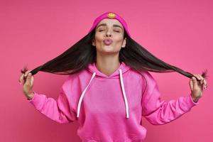 Beautiful woman in hooded shirt holding her long hair and puckering against pink background photo