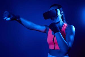 Young woman in virtual reality glasses practicing in punching against colorful background photo