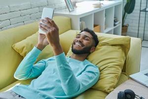 hombre africano feliz usando un teléfono inteligente mientras se relaja en el sofá en casa foto