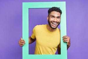 hombre africano feliz mirando a través de un marco de imagen mientras está de pie contra el fondo púrpura foto