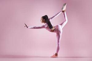 Beautiful young woman in sports clothing doing stretching exercises against pink background photo