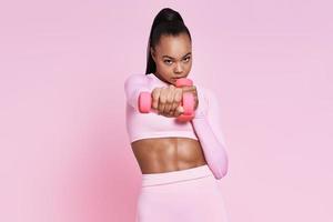 mujer joven concentrada haciendo ejercicio con pesas mientras está de pie contra el fondo rosa foto
