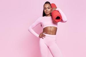 Confident African woman in sports clothing carrying exercise mat against pink background photo