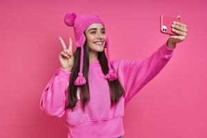 Playful young woman in funky hat making selfie and gesturing against pink background photo