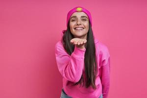 Beautiful young woman in funky hat blowing a kiss against pink background photo