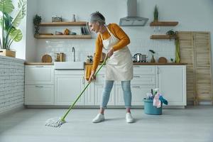 Full length of confident senior woman cleaning floor with mop while standing at the domestic kitchen photo