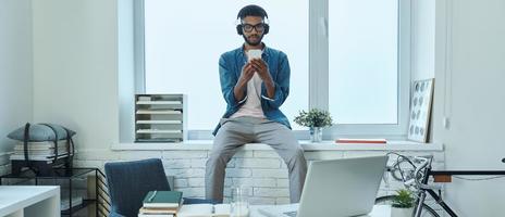 Confident African man using smart phone while sitting on the window sill in office photo