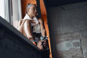 Beautiful woman with towel on shoulders enjoying break while sitting on window sill in gym photo