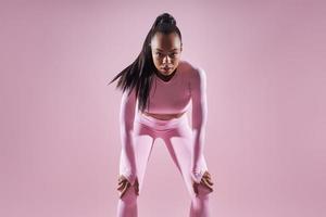 mujer africana concentrada en ropa deportiva cogida de la mano en las rodillas contra fondo rosa foto
