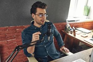 Confident young man using microphone and smiling while recording podcast in studio photo