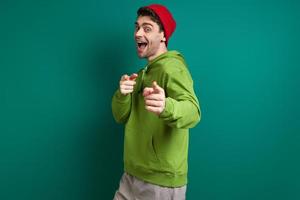 Happy young man pointing camera while standing against green background photo