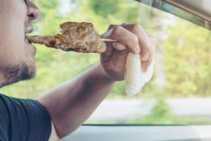 hombre que conduce un automóvil mientras come un palo de cerdo a la parrilla con un palo de bambú de punta afilada y arroz pegajoso en un paquete de plástico peligrosamente - conducción multitarea mala conducta concepto de conducción de riesgo de peligro foto