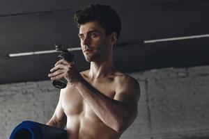 Handsome young man carrying exercise mat and drinking water while standing in gym photo