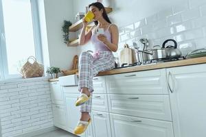 Young woman holding smart phone and drinking coffee while sitting on the kitchen counter photo