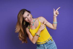 Happy young woman pointing away while standing against purple background photo