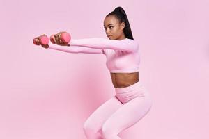 Attractive African woman exercising with dumbbells while standing against pink background photo
