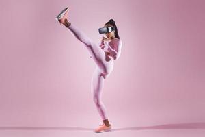 Woman in virtual reality headset practicing in kicking while standing against pink background photo