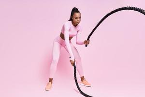 mujer africana joven concentrada haciendo ejercicio con cuerdas de batalla contra fondo rosa foto
