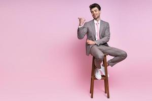 un joven feliz con traje completo señalando mientras se sienta en la silla contra un fondo rosado foto