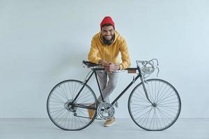 Happy young African man leaning at his bicycle while standing against the wall photo