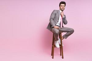 Confident man in full suit adjusting his necktie while sitting on the chair against pink background photo