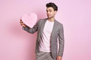 Handsome young man in suit holding paper heart while standing against pink background photo