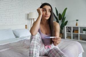 Depressed young woman holding pregnancy test while sitting on bed at home photo