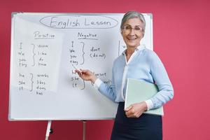 Cheerful senior woman teaching English language near the whiteboard against pink background photo