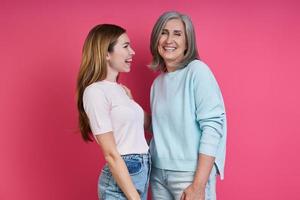 Happy mother and adult daughter standing together against pink background photo