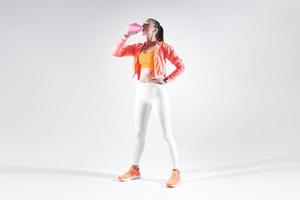 Full length of beautiful young woman in sports clothing drinking water against white background photo