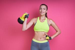 Happy young woman in sports clothing carrying kettlebell against pink background photo