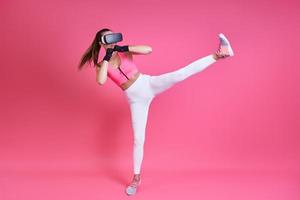 Young woman in virtual reality glasses kicking against pink background photo