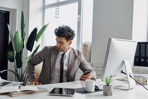 Thoughtful young man holding smart phone while sitting at his working place in office photo