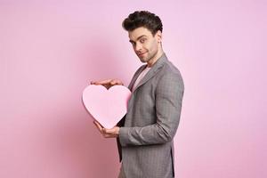 Happy young man in suit holding paper heart while standing against pink background photo