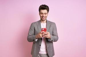 Happy young man in suit using smart phone while standing against pink background photo