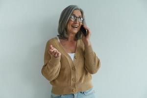 Happy senior woman talking on mobile phone and gesturing while standing against white wall photo