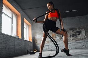 Full length of beautiful African woman carrying battle rope on shoulders while standing in gym photo