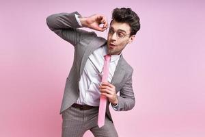 Surprised man in full suit adjusting his eyeglasses while standing against pink background photo