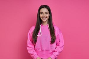 bella joven con camisa encapuchada mirando a la cámara y sonriendo con fondo rosa foto