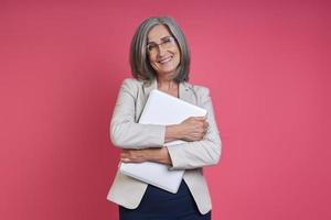 Happy senior woman in formalwear carrying laptop while standing against pink background photo