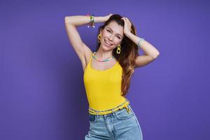 Beautiful young woman holding hands in hair and smiling while standing against purple background photo