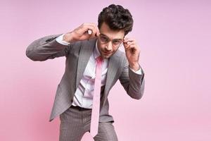 hombre confiado en traje completo ajustando sus anteojos mientras está de pie contra el fondo rosa foto