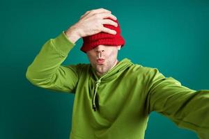 Playful young man making selfie and pulling hat on face while standing against green background photo