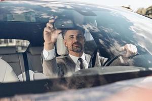 Confident man in formalwear adjusting rear view mirror while sitting on the front seat of a car photo