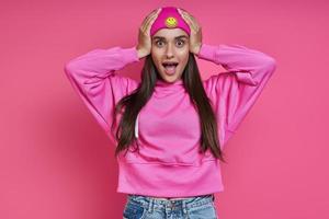 Surprised young woman in hooded shirt holding head in hands against pink background photo