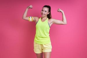 Happy woman in sports clothing wearing captain band and showing biceps against pink background photo