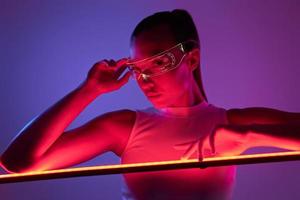 Beautiful woman in futuristic glasses leaning at the LED lamp against dark background photo