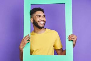 Cheerful African man looking through a picture frame while standing against purple background photo