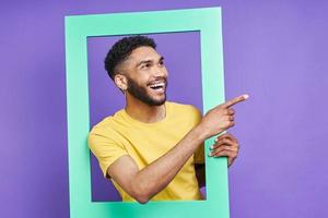 Happy African man looking through a picture frame and pointing away against purple background photo