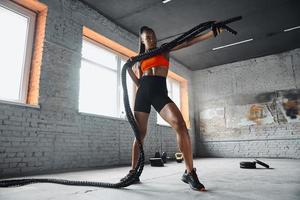 Beautiful young African woman examining battle rope while standing in gym photo